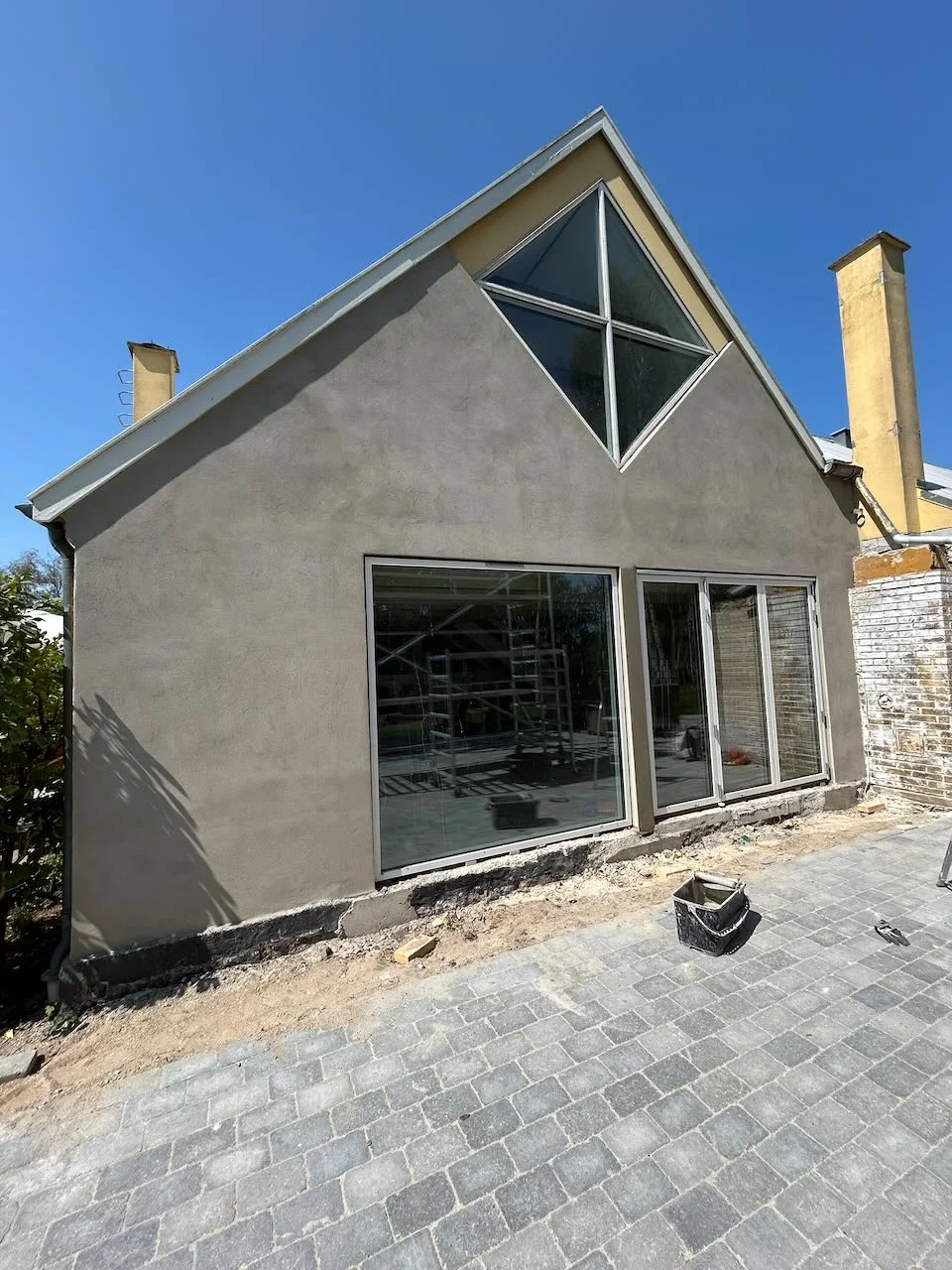 Et moderne hus med frisk puds, store vinduer og et trekantet vindue øverst er et vidnesbyrd om en professionel boligrenovering. Fasaden er endnu ikke færdig, og i forgrunden ses byggematerialer og brosten under en klarblå himmel – et bevis på det kvalitetshåndværk, som Tømrerfirmaet Kenn leverer.