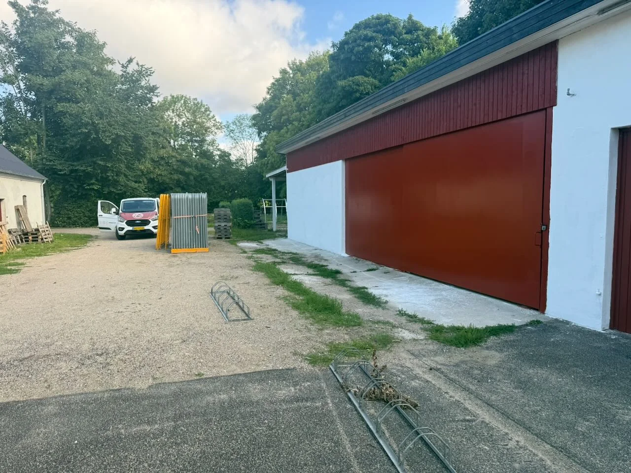 En stor rød og hvid bygning, muligvis stedet for en boligrenovering udført af Tømrerfirmaet Kenn, ligger ved siden af en grusindkørsel. En hvid varevogn holder parkeret tæt på nogle stablede metalhegn. Området er omgivet af træer og græs, og himlen er delvist overskyet.