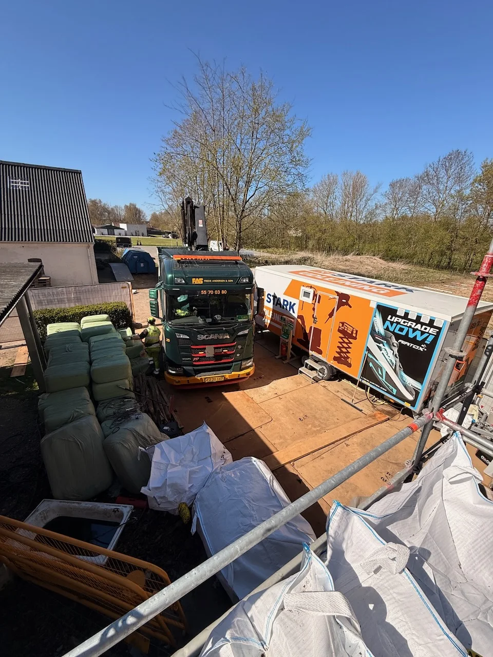 En grøn lastbil og en orange påhængsvogn fra Tømrerfirmaet Kenn står parkeret på en byggeplads ved siden af byggematerialer, indpakkede paller og udstyr under en klar blå himmel med træer og bygninger i baggrunden, klar til tømrer- og renoveringsprojekter.