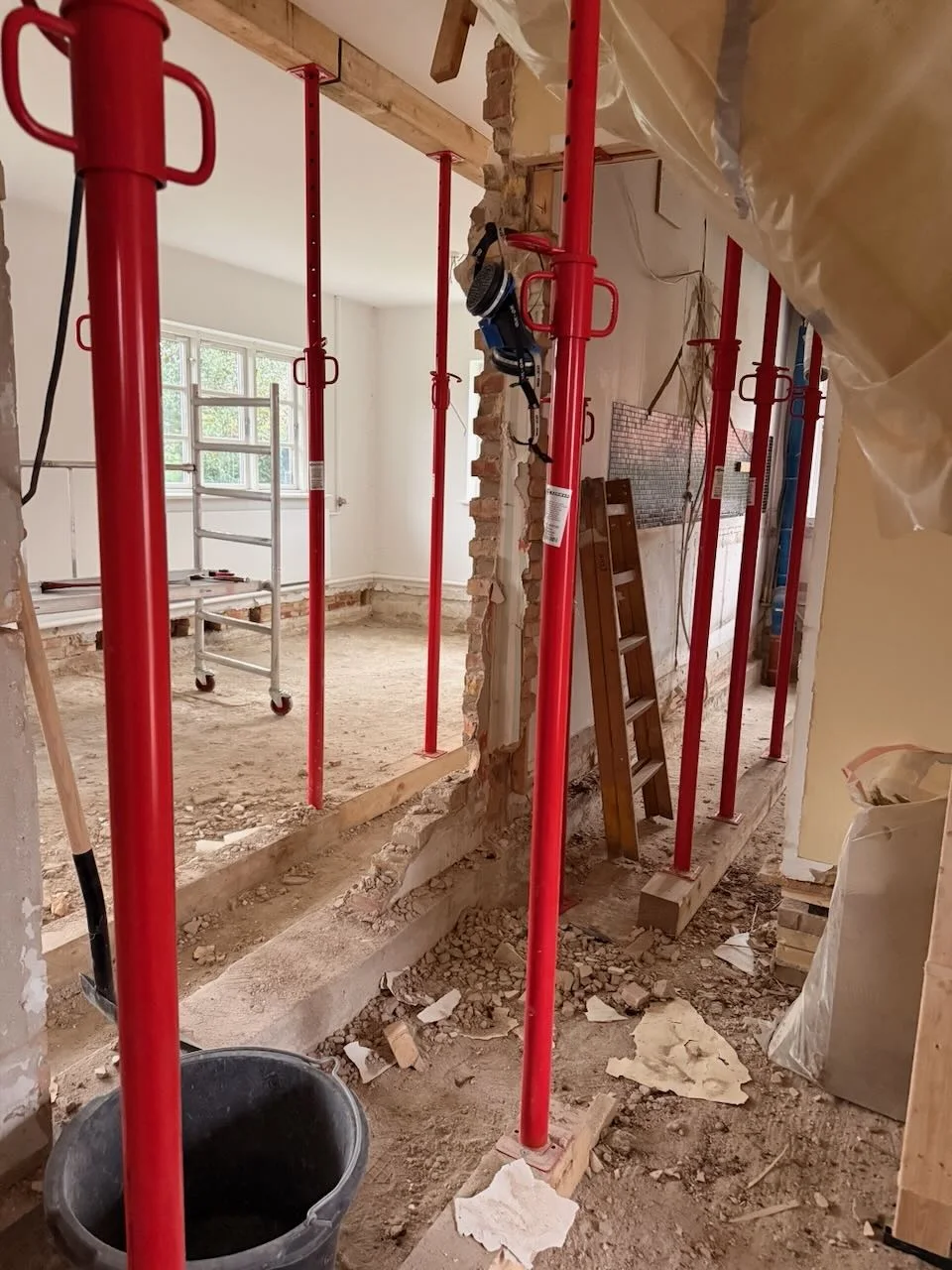 Et rum under renovering med røde støttepæle, synlige vægstolper, affald på gulvet, en sort spand, værktøj og en stige. Sollys strømmer ind gennem vinduerne i baggrunden – et tydeligt tegn på, at Tømrerfirmaet Kenn er i gang med tømrerarbejdet i forbindelse med dette boligrenoveringsprojekt.