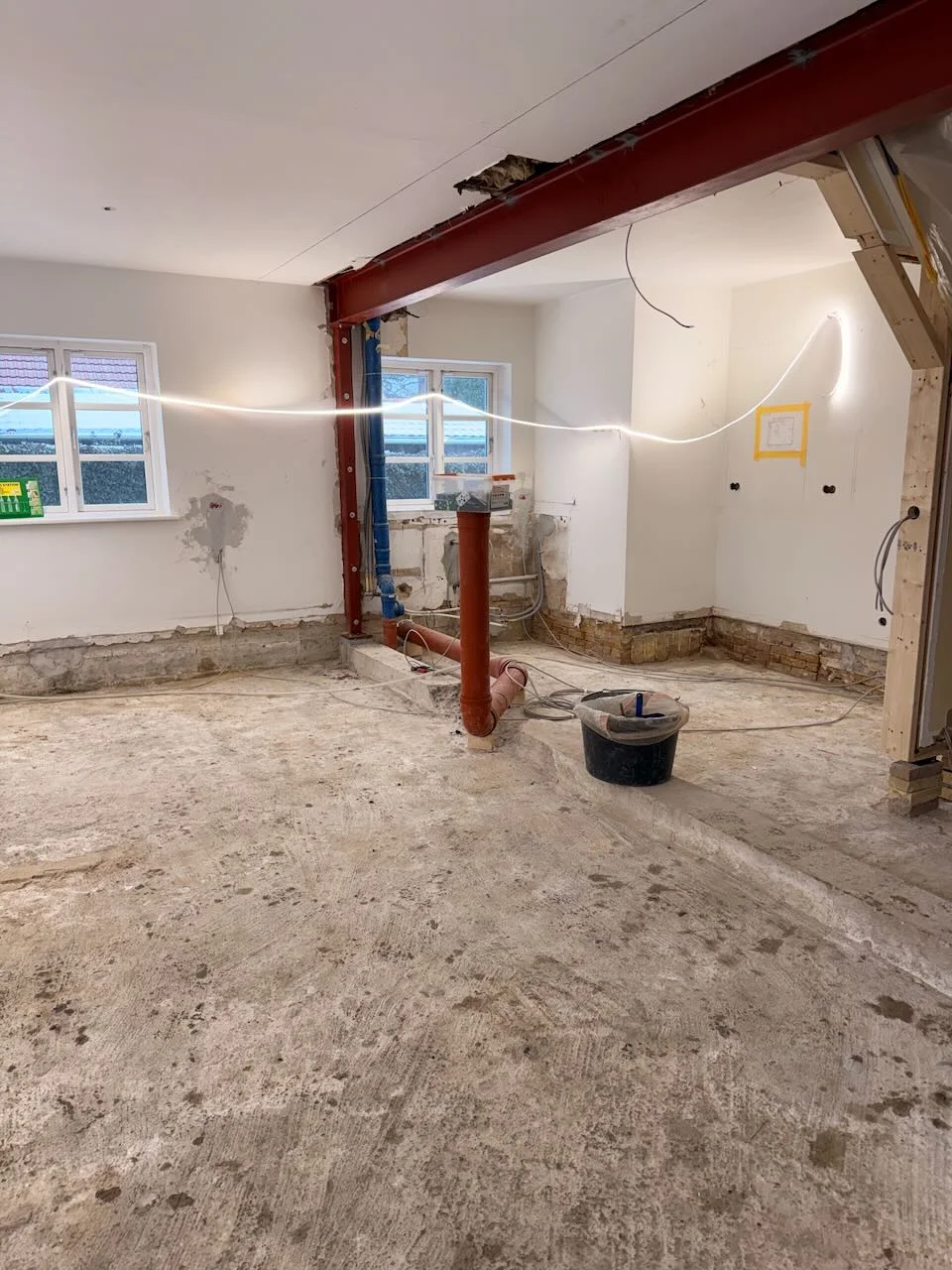 I et rum, der er ved at blive renoveret af Tømrerfirmaet Kenn, ses synlige rør, ledninger og en stålbjælke. Væggene er delvist afklædt, gulvet er af rå beton, og der ligger tømrerværktøj spredt rundt omkring. To vinduer sørger for naturligt lys.