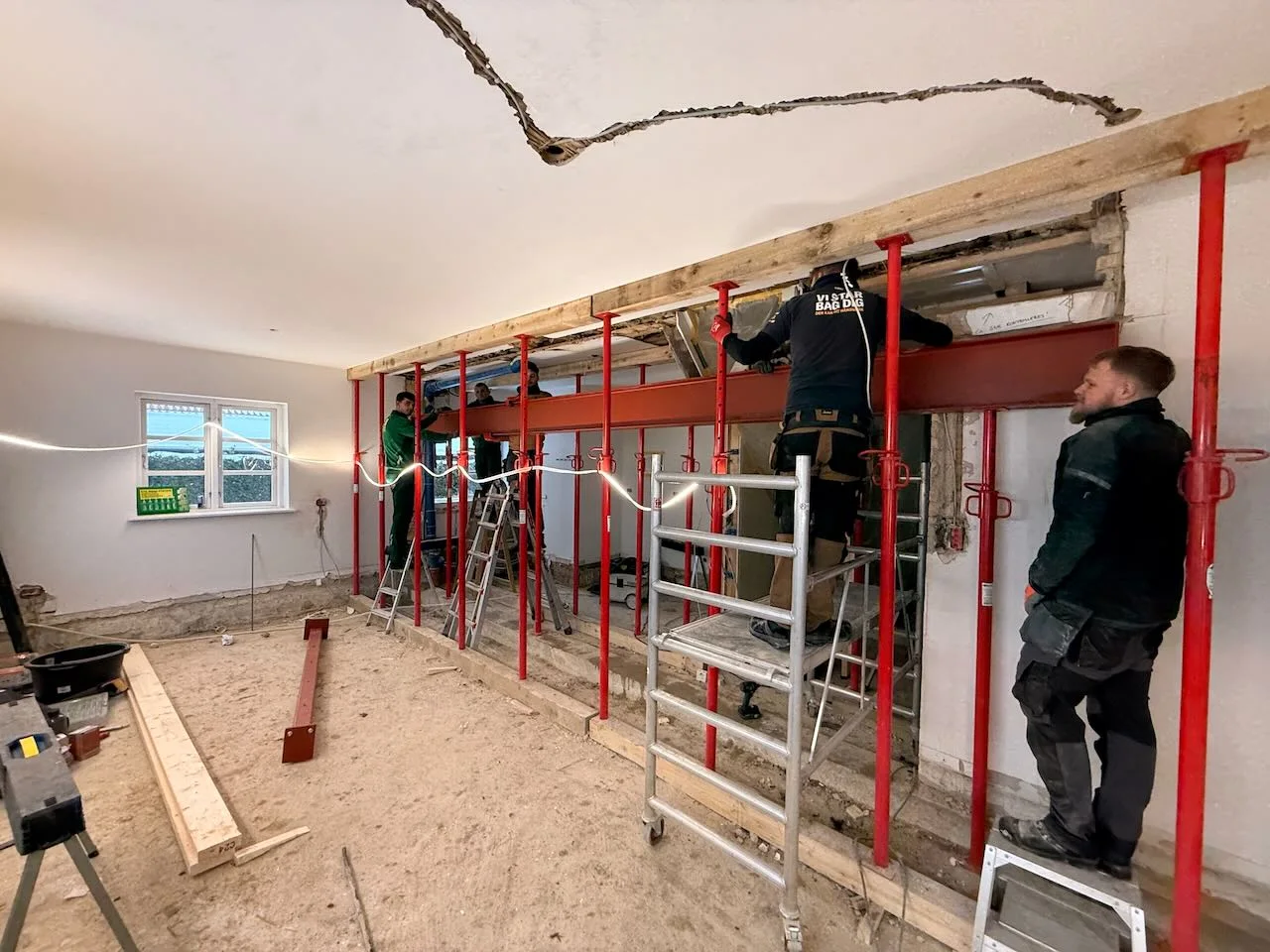 Fire medarbejdere fra Tømrerfirmaet Kenn monterer en stålbjælke indendørs ved hjælp af røde støttepæle og stilladser. Loftet, der viser tegn på slitage med en stor revne, vidner om den omfattende renovering af huset, der er i gang. Byggeværktøj og materialer ligger spredt på gulvet, mens et lille vindue lukker dagslys ind og oplyser den travle tømrerværkplads.
