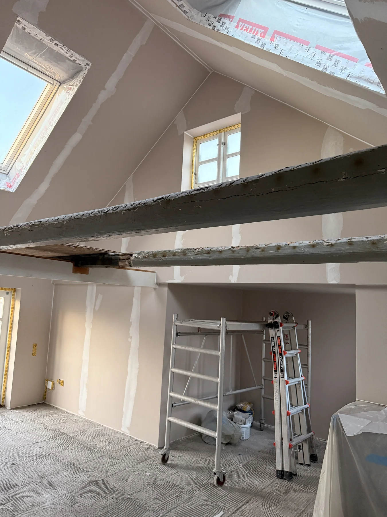 Et rum, der er under renovering, med synlige bjælker, et metalstillads, en trappestige og synlige pudsefuger. Tagvinduer og et lille vindue sørger for naturligt lys, mens gulvet er dækket af beskyttelsesmateriale – et tydeligt bevis på det dygtige tømrerarbejde udført af Tømrerfirmaet Kenn.