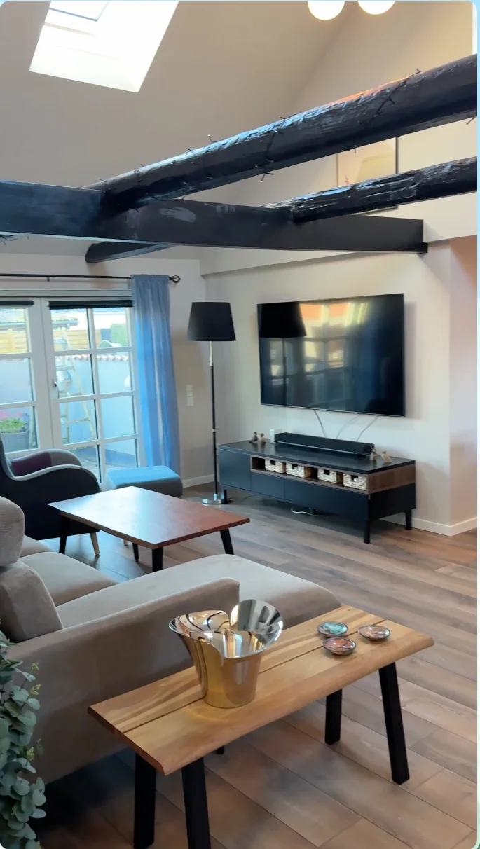 En moderne stue med synlige mørke loftsbjælker, der vidner om mesterligt tømrerhåndværk, et stort vægmonteret tv, en sofa, et sofabord i træ, en gulvlampe og et lille bord med service. Der er store vinduer med blå gardiner og trægulv – et indbydende rum, der er perfekt til ethvert boligrenoveringsprojekt udført af Tømrerfirmaet Kenn.