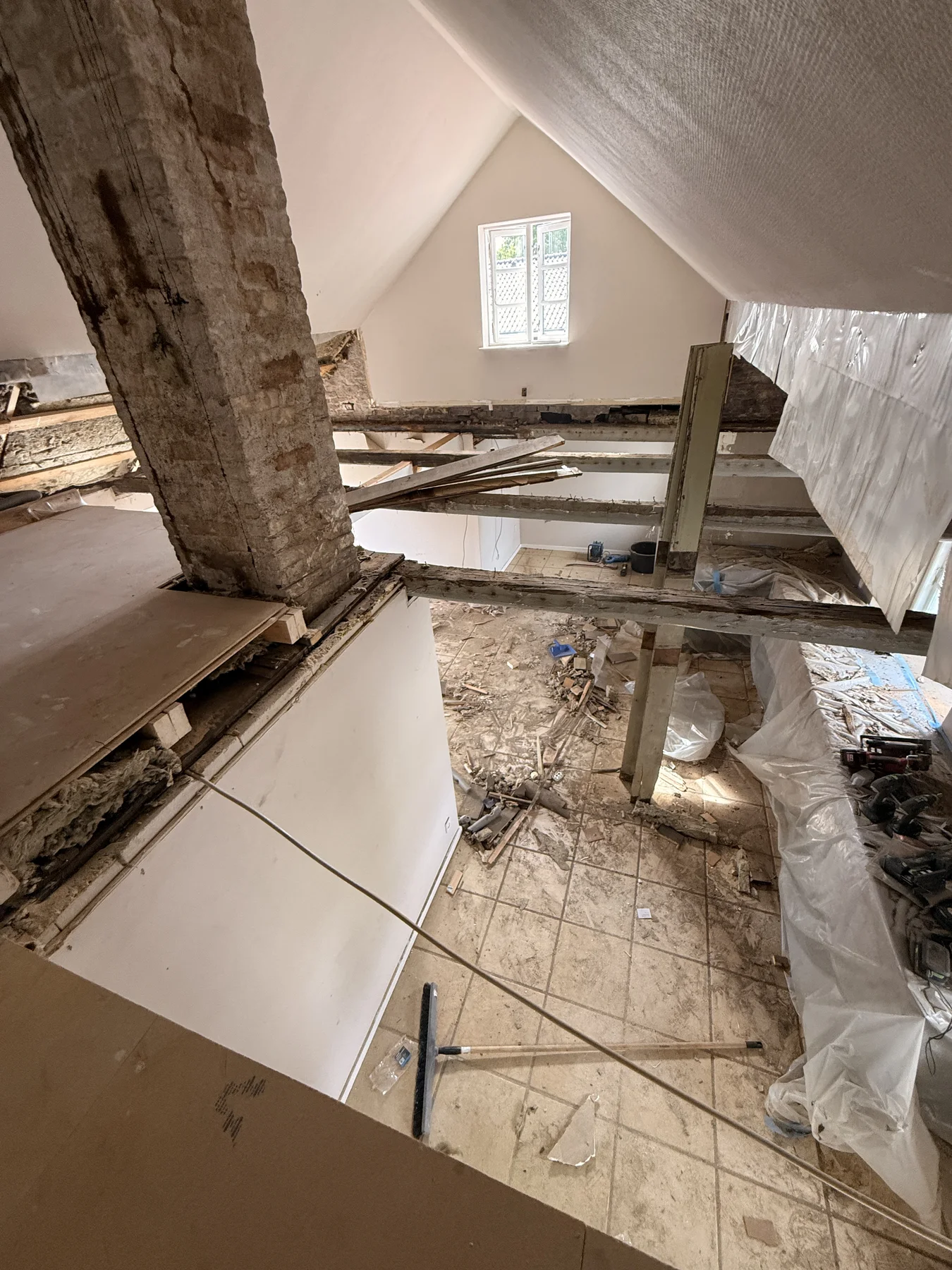 Udsigt til et hus under renovering med synlige bjælker, byggeaffald på klinkegulvet, et delvist fjernet loft og byggematerialer spredt overalt. Det dygtige tømrerarbejde udført af Tømrerfirmaet Kenn træder tydeligt frem, når lyset strømmer ind gennem et lille vindue i den fjerne ende af rummet.