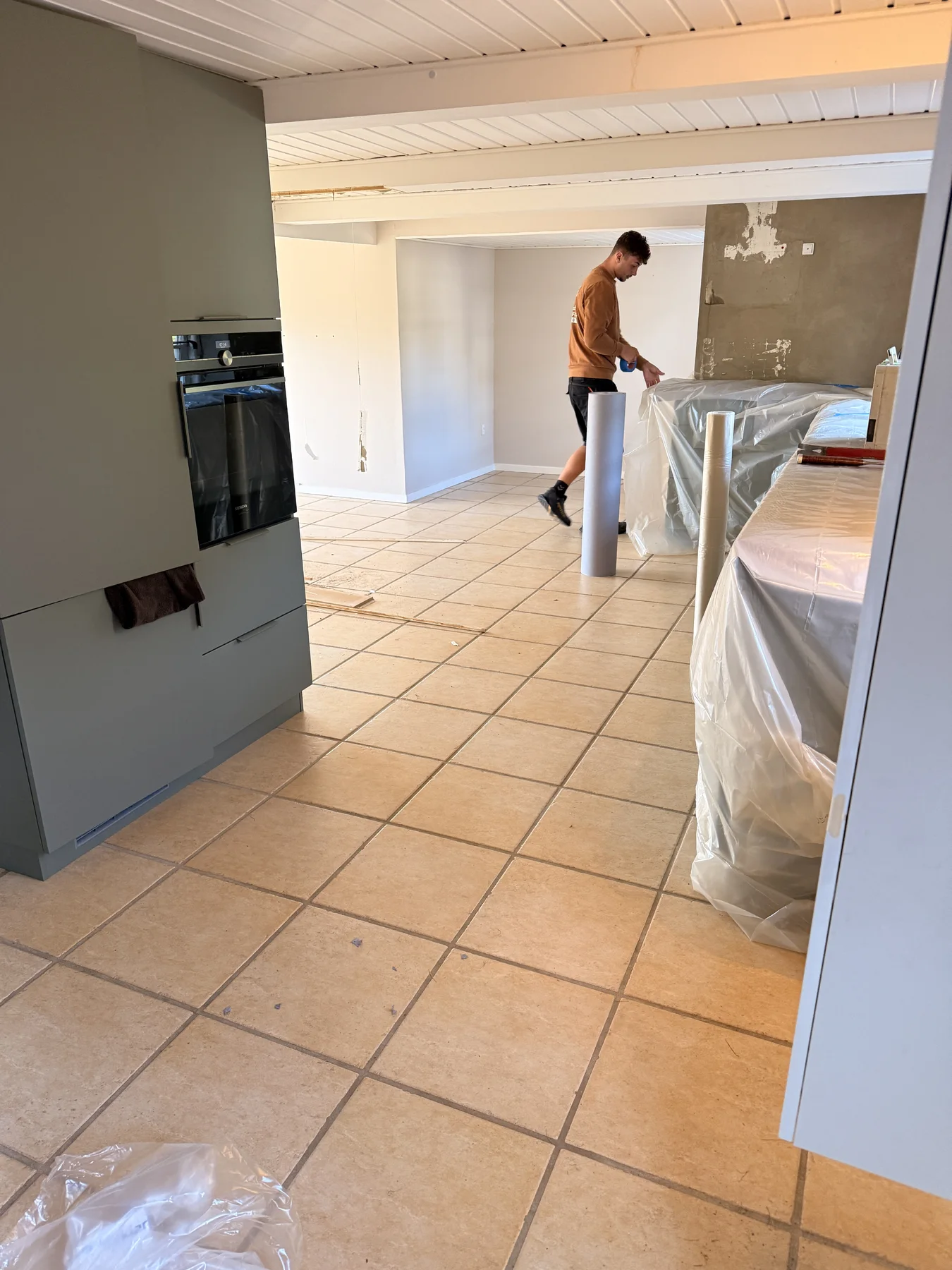 En person står i et køkken, der er under renovering, og holder en rulle med materiale i hånden. Hvidevarer og overflader er dækket af plastfolie. Væggene bærer præg af det igangværende arbejde, hvilket vidner om det dygtige tømrerhåndværk, der er lagt for dagen, og gulvet er flisebelagt. Rummet fremstår rent og velordnet, hvilket afspejler den professionalisme, der er karakteristisk for Tømrerfirmaet Kenn i forbindelse med boligrenoveringer.