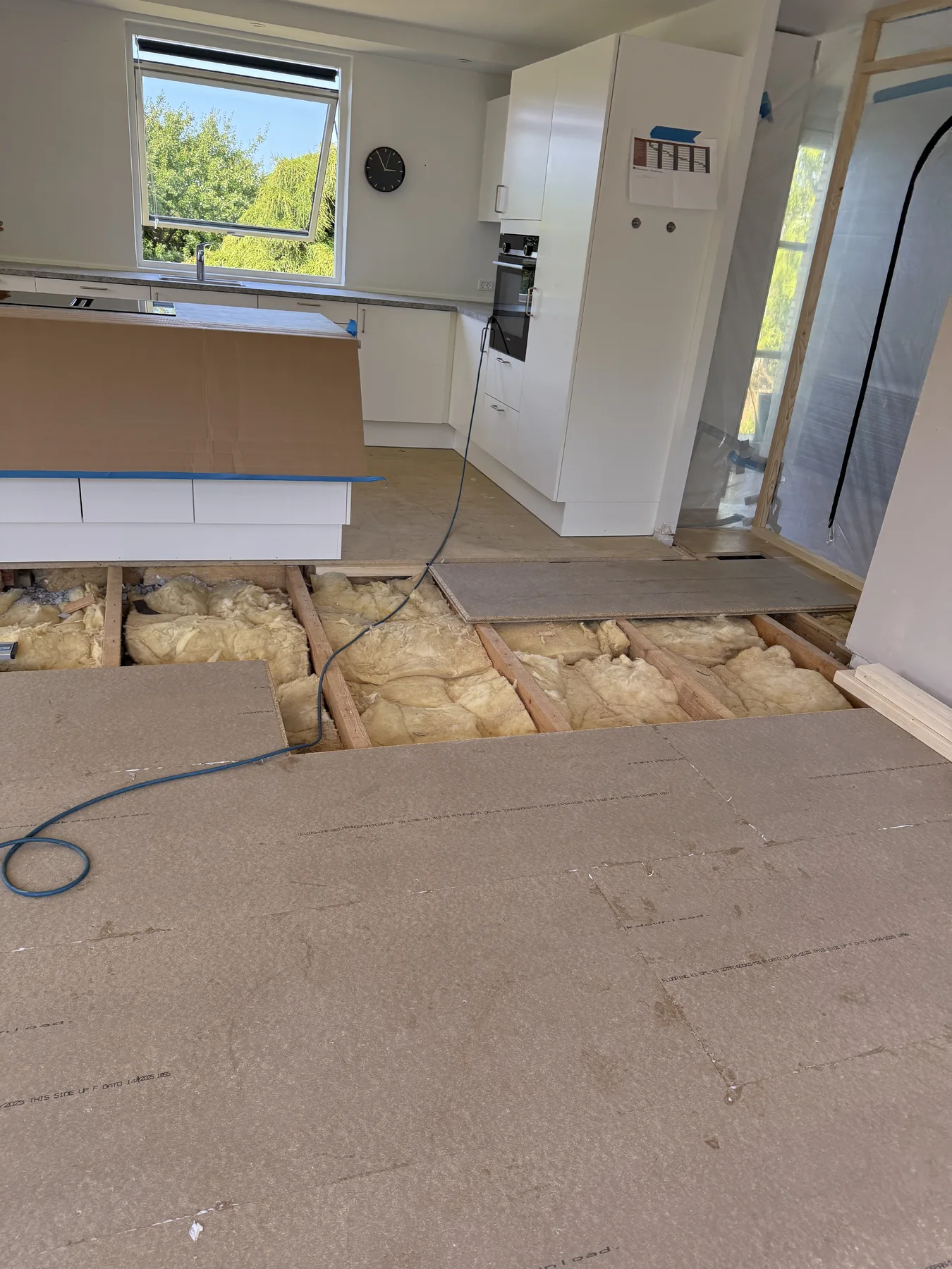 Et køkken under renovering, hvor en del af gulvet er fjernet, så isoleringen og bjælkerne er synlige – et tydeligt tegn på, at der er dygtige tømrere i gang med arbejdet. Skabe og hvidevarer er monteret, og midt i rummet står en køkkenø, der er dækket af pap. Gennem vinduet kan man se grønne omgivelser udenfor, hvilket understreger den forvandling, som Tømrerfirmaet Kenns boligrenoveringsteam er i gang med.