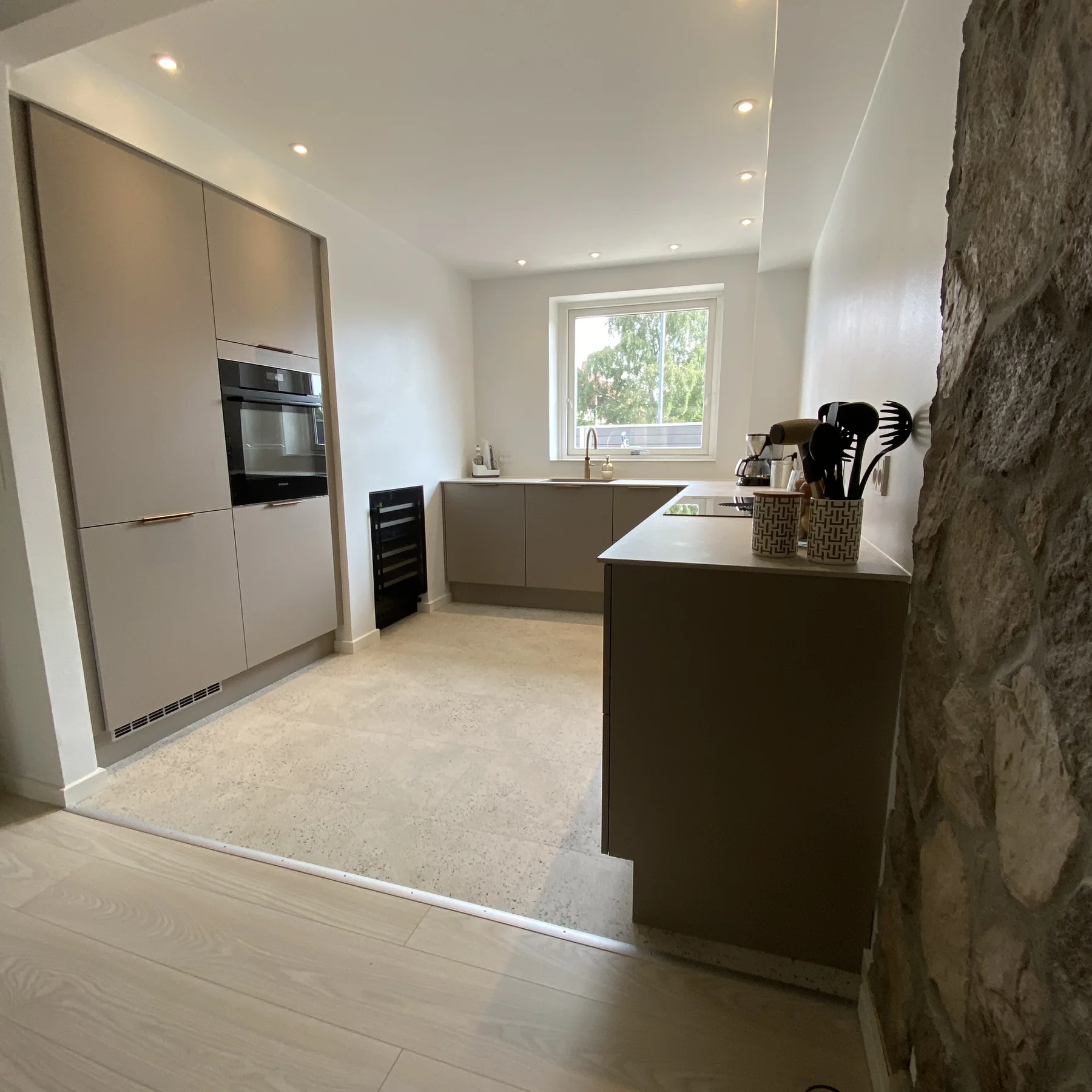 Moderne køkken med beige skabe, indbygget ovn, sort vinkøler, bordmodeller, et vindue med udsigt til det fri og en delvis stenvæg til højre. Tømrerfirmaet Kenns professionelle håndværk kommer tydeligt til udtryk i hele rummet og suppleres af indbyggede loftslamper og lyse gulve, hvilket skaber en frisk stemning efter renoveringen.