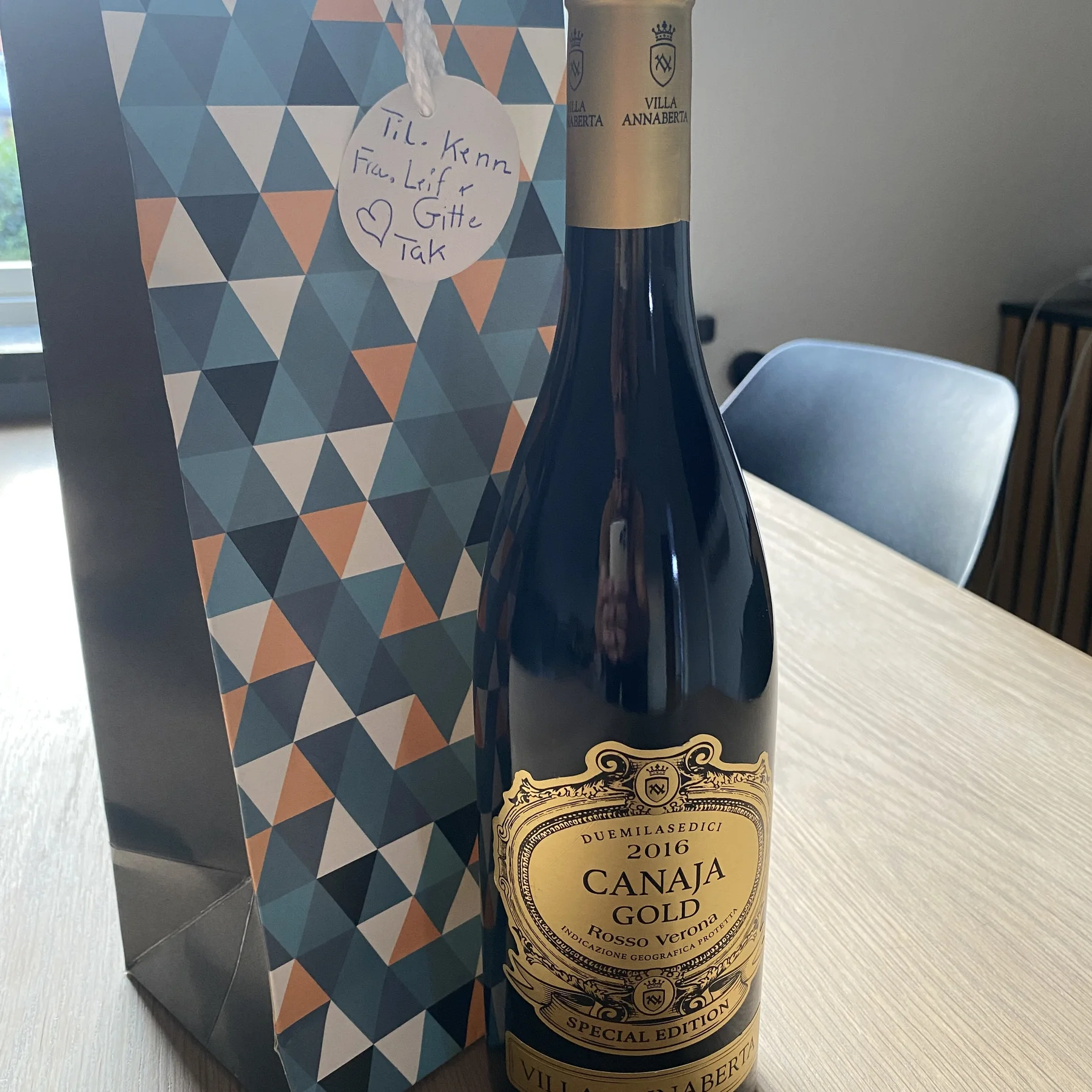 En flaske Canaja Gold-vin fra 2016 står på et bord ved siden af en mønstret gavepose med et hjerteformet kort med håndskrevet tekst.