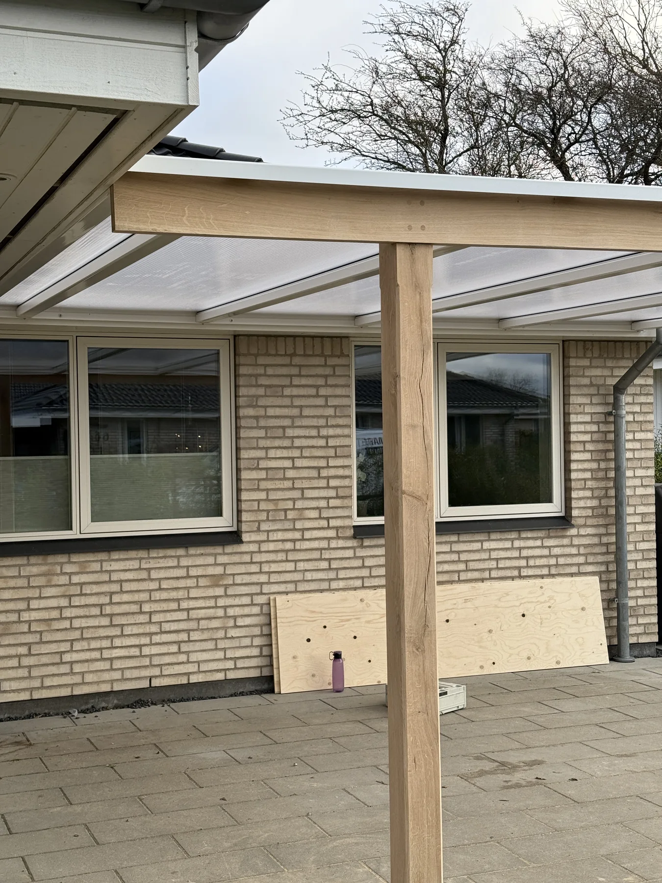 En træpæl bærer et delvist opført tag over en terrasse, der er fastgjort til et murstenshus, hvilket viser, at renoveringen af huset er i gang. Man kan se tre vinduer, og en krydsfinerplade – som er uundværlig i tømrerprojekter – ligger op ad væggen på en belagt overflade, hvilket vidner om det dygtige håndværk hos Tømrerfirmaet Kenn.
