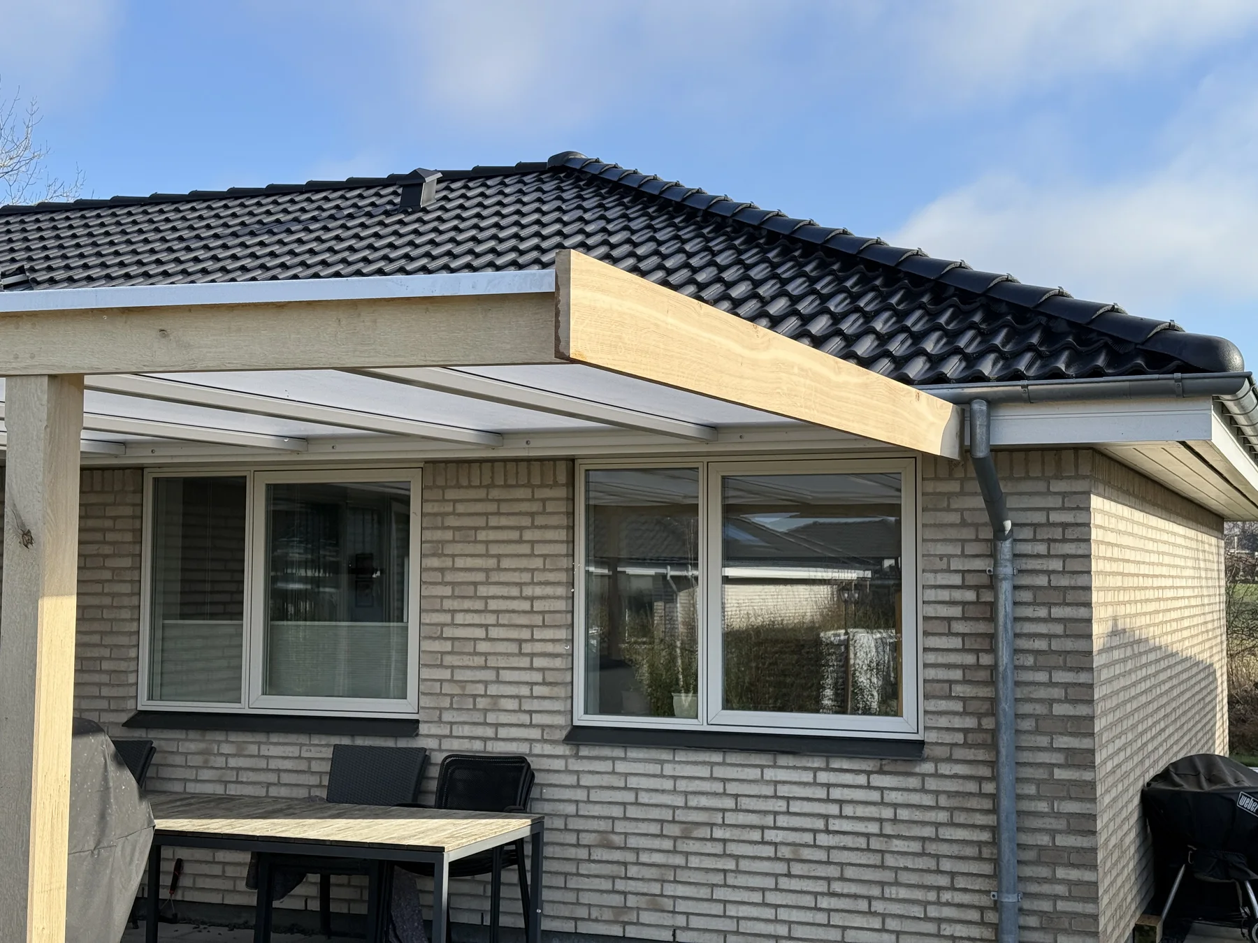 Et murstenshus med tre vinduer, et mørkt tegltag og en overdækket terrasse med træramme og gennemsigtigt tag – mesterligt udført af Tømrerfirmaet Kenn, der er specialister i tømrerarbejde og boligrenovering. Der står et bord og stole på terrassen.