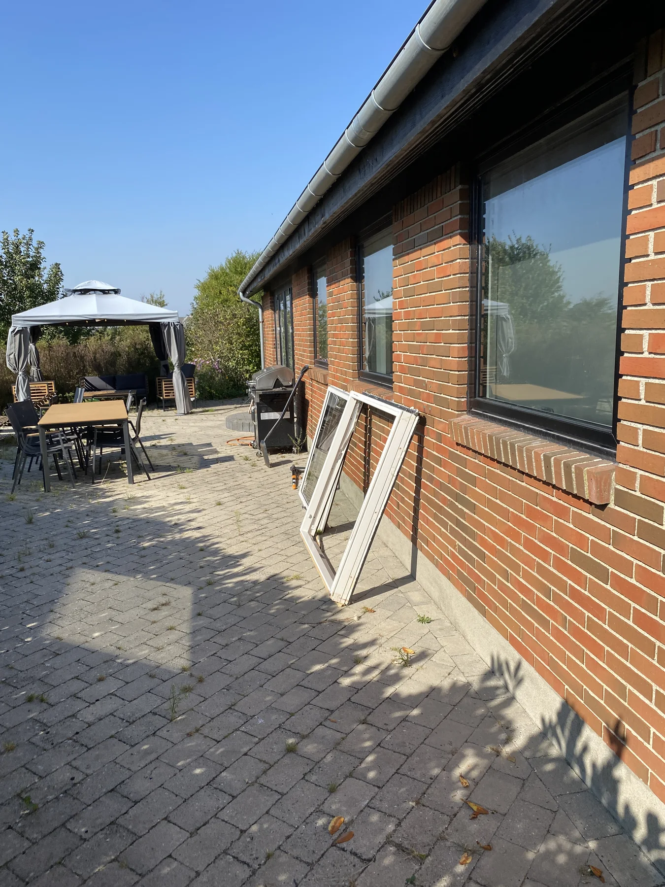 Et rødt murstenshus med store vinduer har to vinduesrammer, der står op ad væggen, hvilket tyder på, at Tømrerfirmaet Kenn er i gang med tømrerarbejde eller renovering af huset. På terrassen står der et bord med stole under en baldakin, og der er en grill i nærheden. I baggrunden ses buske og træer.