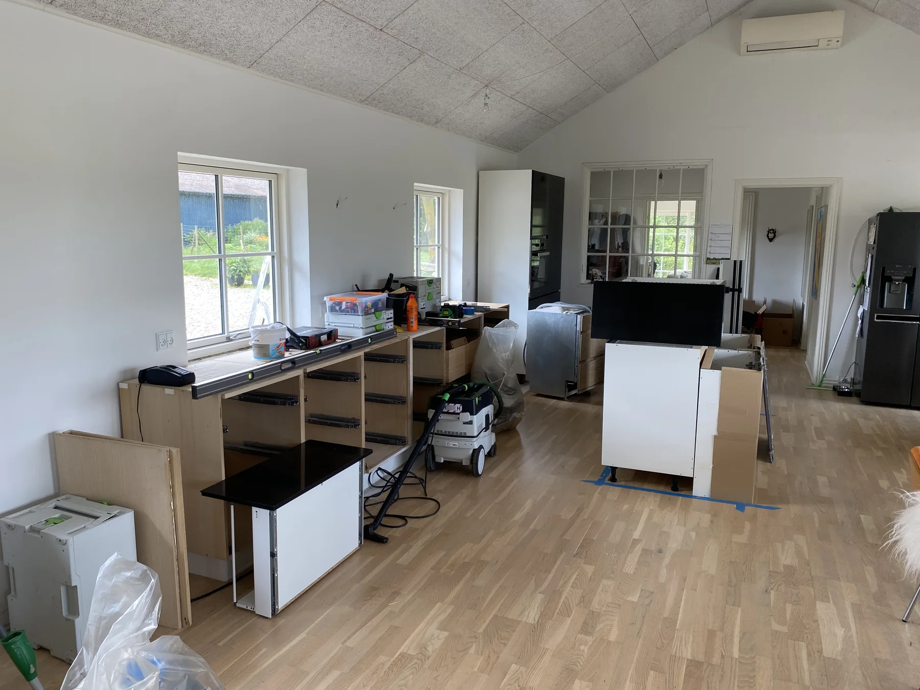 Et køkken under renovering, hvor skabe og skuffer er delvist monteret, værktøj og materialer ligger på gulvet, og hvidevarer ses i baggrunden, afspejler den igangværende renovering af boligen. Det lyse trægulv og de hvide vægge er tydelige og fremhæver det professionelle tømrerarbejde fra Tømrerfirmaet Kenn.