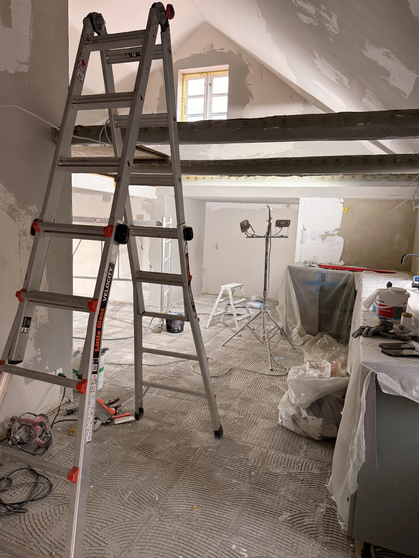 En metalstige står midt i et rum, der er under renovering, hvilket vidner om, at Tømrerfirmaet Kenn er i gang med at renovere boligen. Værktøj, lamper og tildækkede møbler ligger spredt rundt omkring, mens tømrerarbejdet skrider frem. Væggene er delvist pudset, og naturligt lys strømmer ind gennem et lille vindue højt oppe.