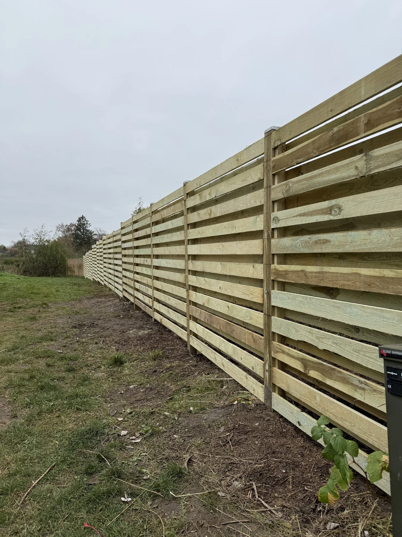 Et nyopført træhegn, udført af Tømrerfirmaet Kenn, strækker sig langs kanten af en græsplæne under en overskyet himmel. Hegnet består af vandrette planker og solide lodrette stolper – et bevis på tømrerarbejde af høj kvalitet. I forgrunden ses nogle planter og et lille grønt område, der tilfører dette nylige boligrenoveringsprojekt et friskt præg.