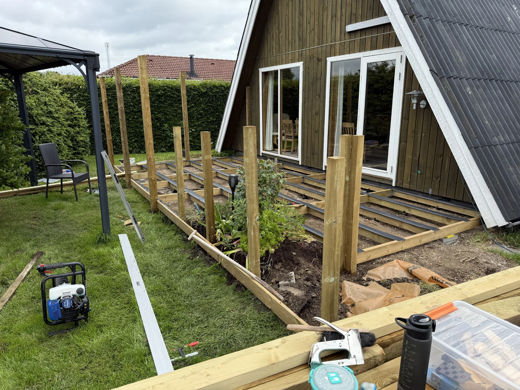 Der er gang i opførelsen af en træterrasse uden for et hus med glasskydedøre, under tilsyn af Tømrerfirmaet Kenn. De lodrette støttepæle er sat på plads, og terrassen er indrammet af tømmer som en del af et større renoveringsprojekt af huset. Værktøj og materialer ligger spredt i nærheden på græsset og træbjælkerne, hvilket understreger det dygtige tømrerarbejde, der er i gang.