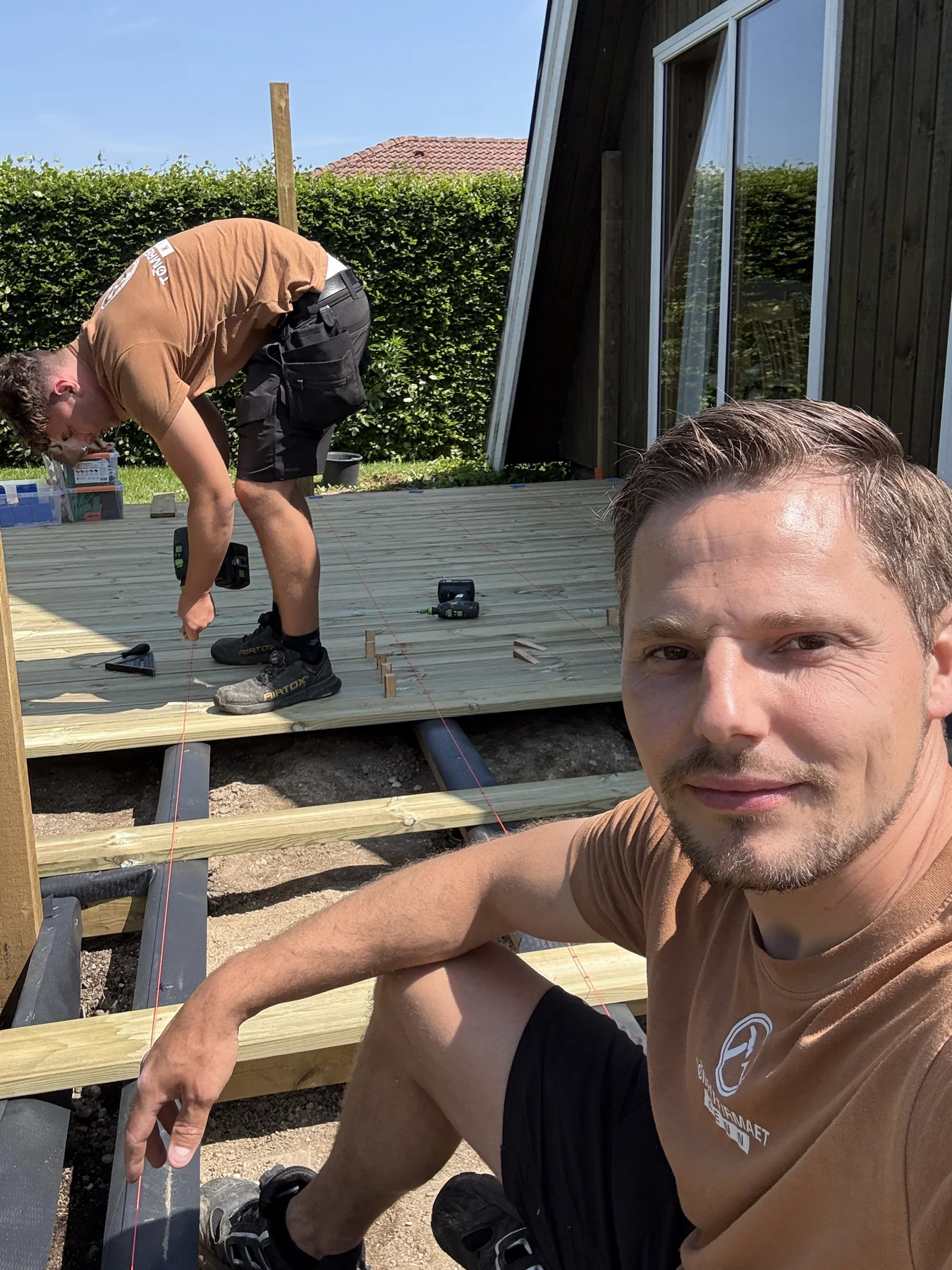 To mænd fra Tømrerfirmaet Kenn er i gang med at bygge en træterrasse udendørs. Den ene mand sidder på knæ og tager et selfie, mens den anden bruger en boremaskine på terrassen som led i deres tømrerprojekt. Der ligger forskellige værktøjer spredt ud over overfladen. Begge har brune skjorter og sorte shorts på.