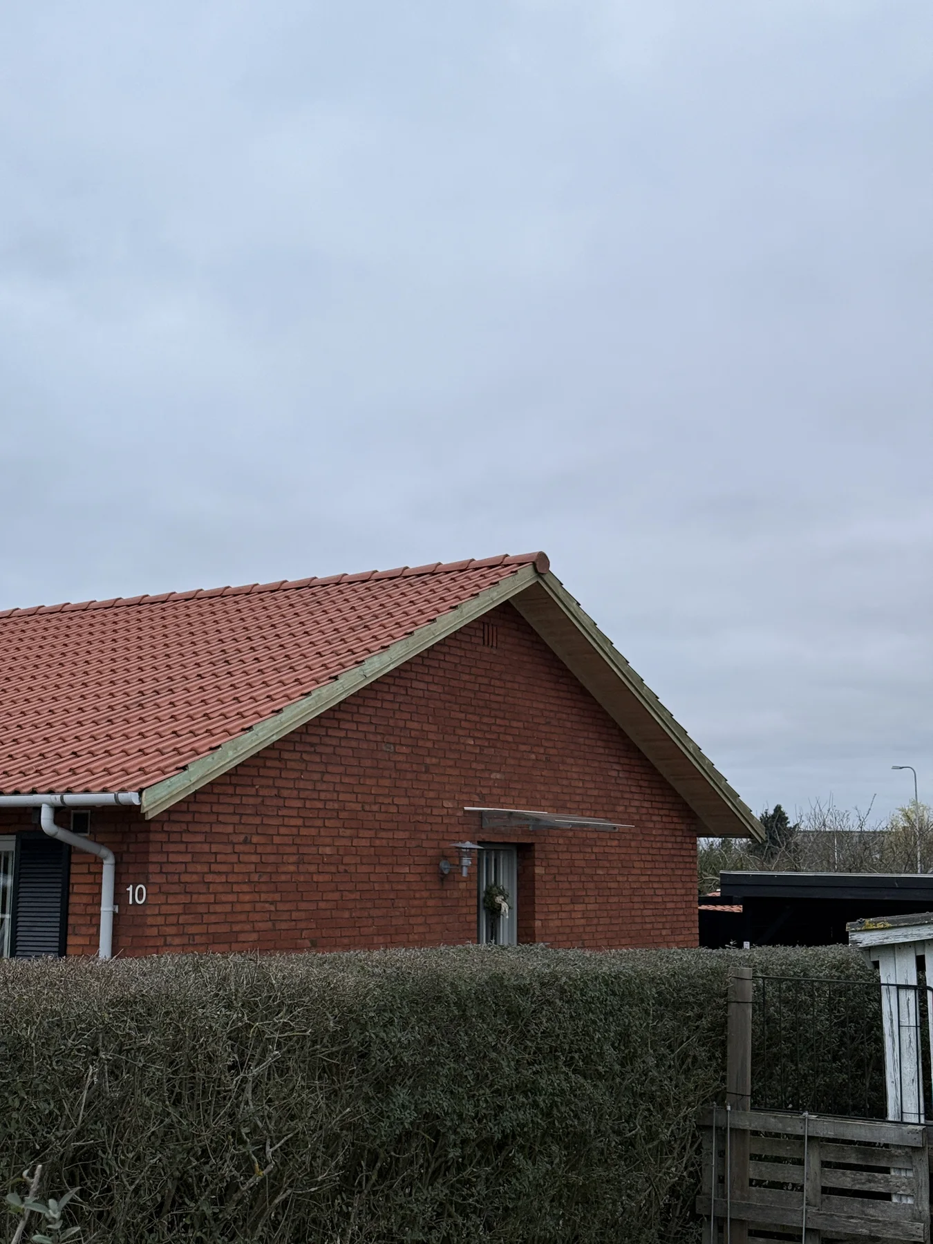 Et hus af røde mursten med rødt tegltag og en hæk foran, som Tømrerfirmaet Kenn har vedligeholdt med stor omhu. Himlen er overskyet. Tallet 10 kan ses på husets væg tæt ved indgangen, hvilket vidner om, at der for nylig er blevet udført renoveringsarbejde på huset. Til højre ses en del af en indkørsel og et hvidt hegn.