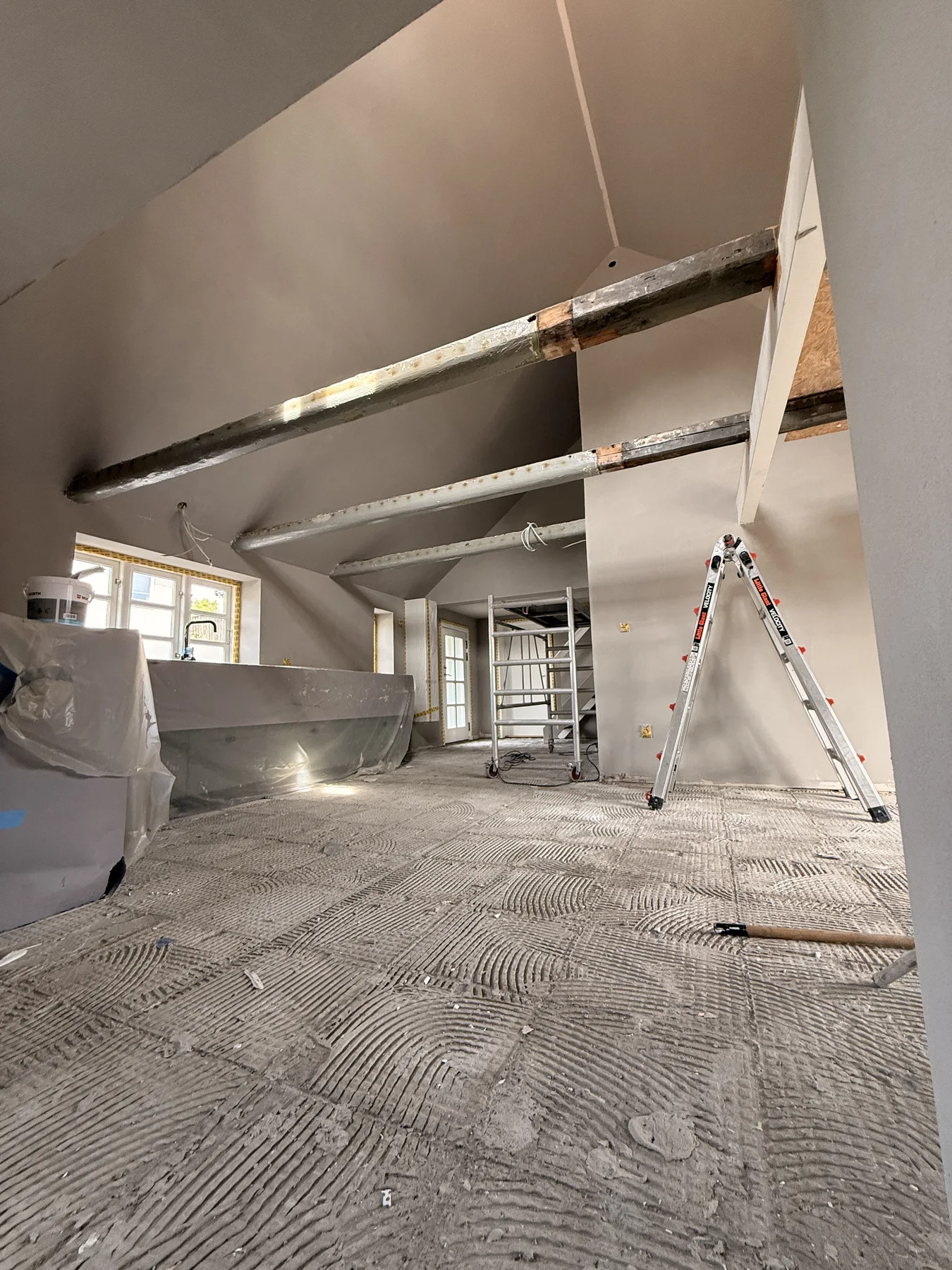 Et rum under renovering med synlige bjælker, møbler dækket af plast, en stige og et stillads. Gulvet ser ufærdigt ud, hvilket afspejler det igangværende tømrerarbejde, der udføres af Tømrerfirmaet Kenn, mens lyset strømmer ind gennem flere vinduer på venstre væg.