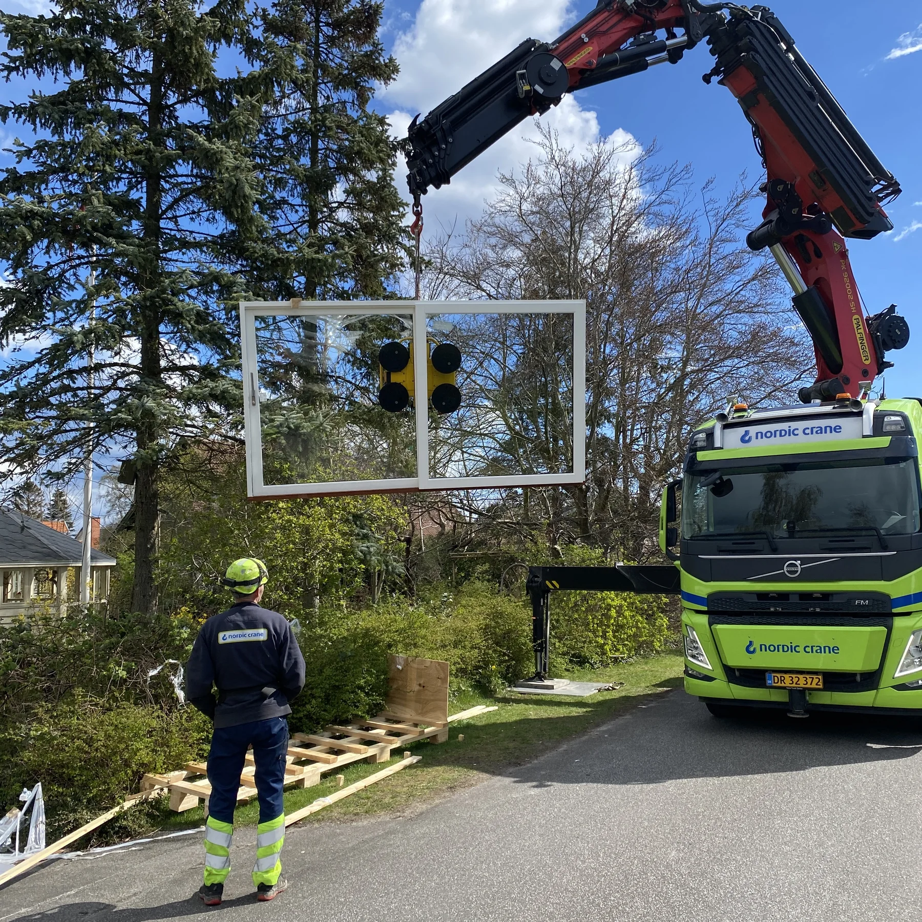 En medarbejder i sikkerhedsudstyr ser til, mens en kran monteret på en Nordic Crane-lastbil løfter en stor vinduesramme ved siden af træer og beboelseshuse på en solrig dag, hvilket viser præcisionen og ekspertisen inden for tømrerfaget i forbindelse med et boligrenoveringsprojekt udført af Tømrerfirmaet Kenn.