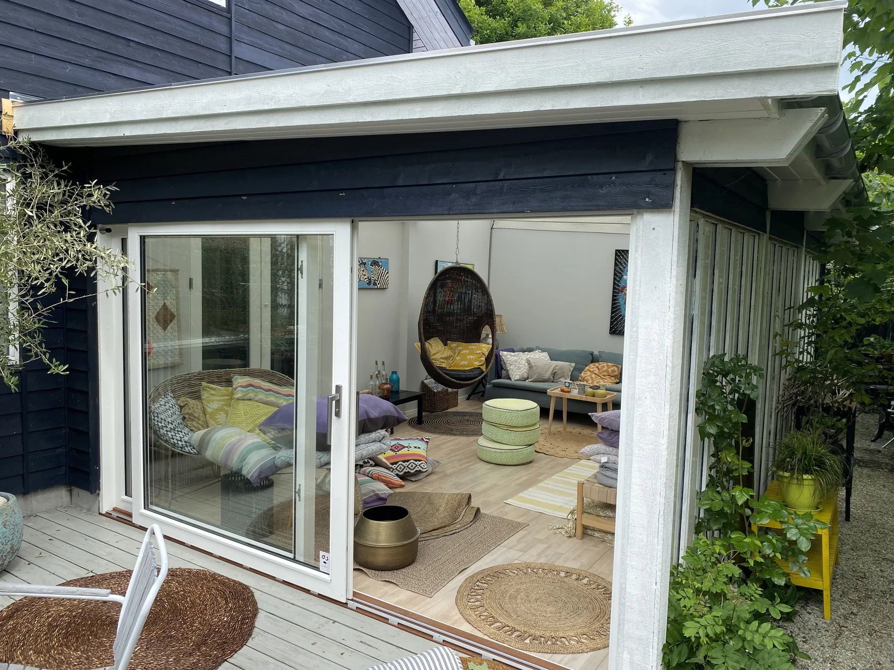 En hyggelig indendørs gårdhave med store glasdøre, farverige puder, en hængestol, runde gulvmåtter og et lille bord. Det er tydeligt, at der er lagt stor vægt på tømrerarbejdet i hele rummet, især i det specialfremstillede træværk, der indrammer gårdhaven. Indretningen omfatter tæpper, puder og stueplanter, hvilket skaber en afslappet og indbydende atmosfære, der er perfekt til ethvert boligrenoveringsprojekt.