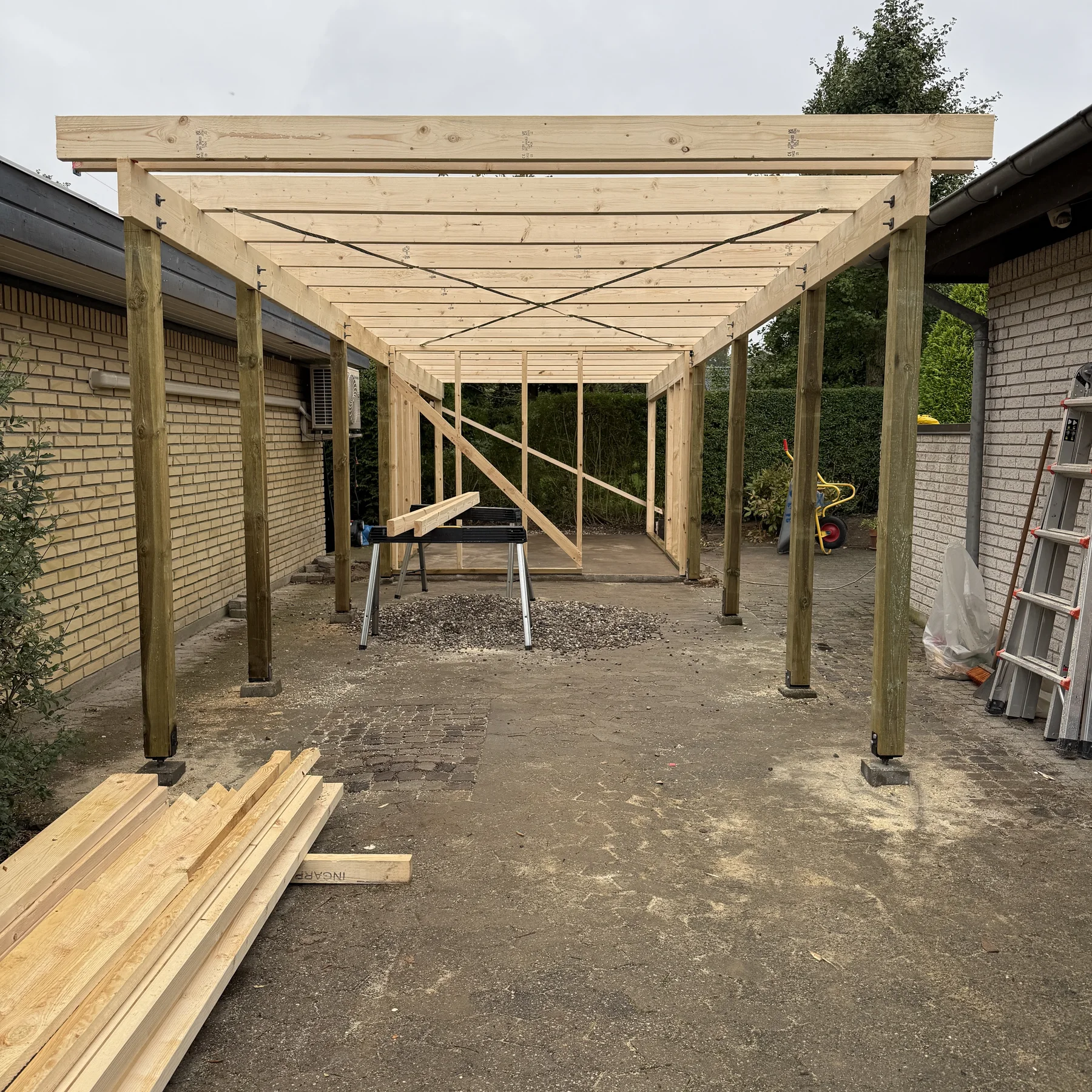Mellem to murstensbygninger er man i gang med at opføre en carport eller et skur af træ, hvilket vidner om det dygtige tømrerarbejde fra Tømrerfirmaet Kenn. Foran bygningen ligger der træbjælker stablet på jorden, og rundt om byggepladsen kan man se værktøj, et savbord og en stige – et livligt billede af en igangværende renovering af et hus.