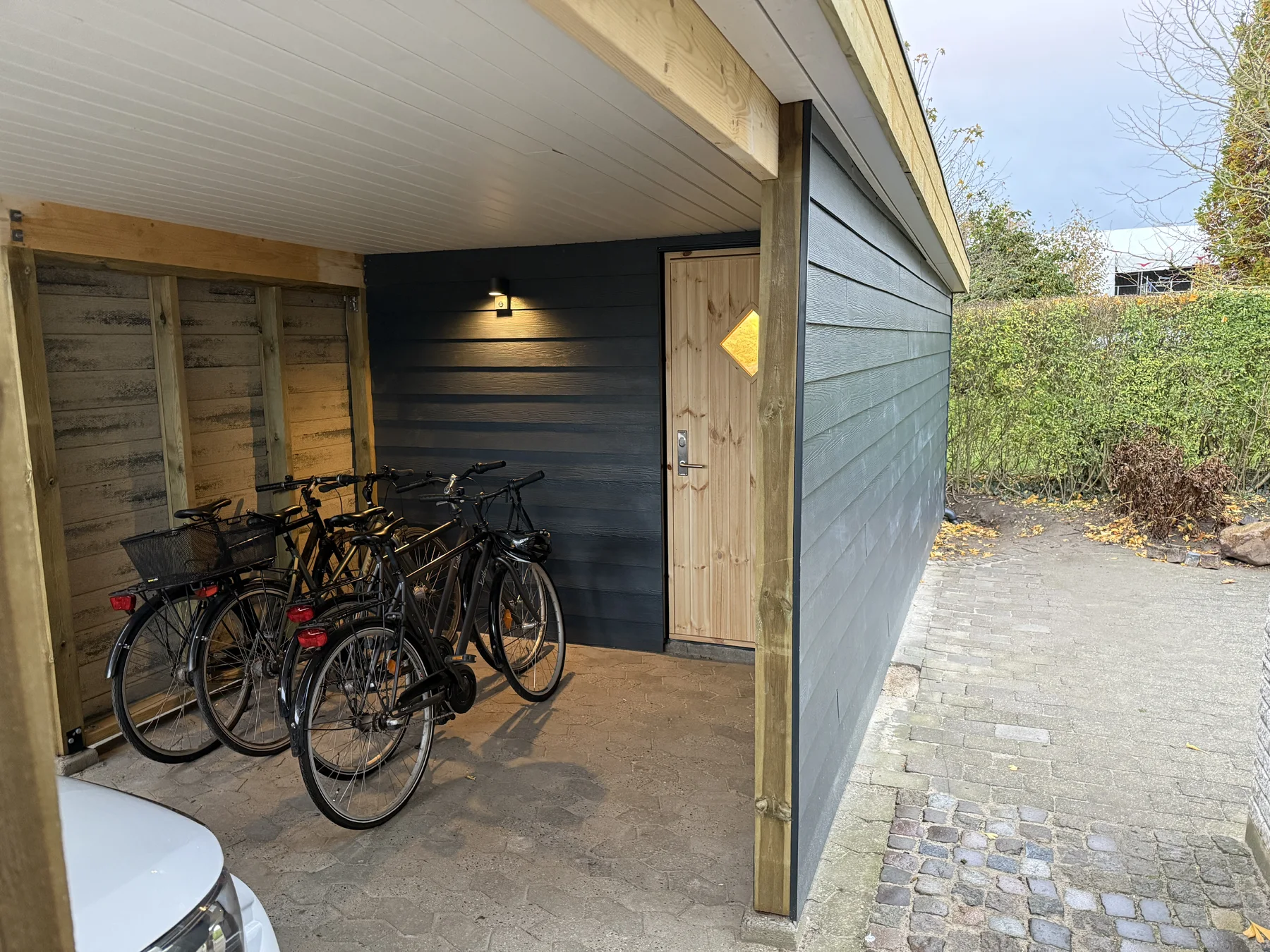 Fire cykler står under en carport ved en mørk træbeklædt væg med trædør og lampe. En bil og hæk ses i baggrunden.