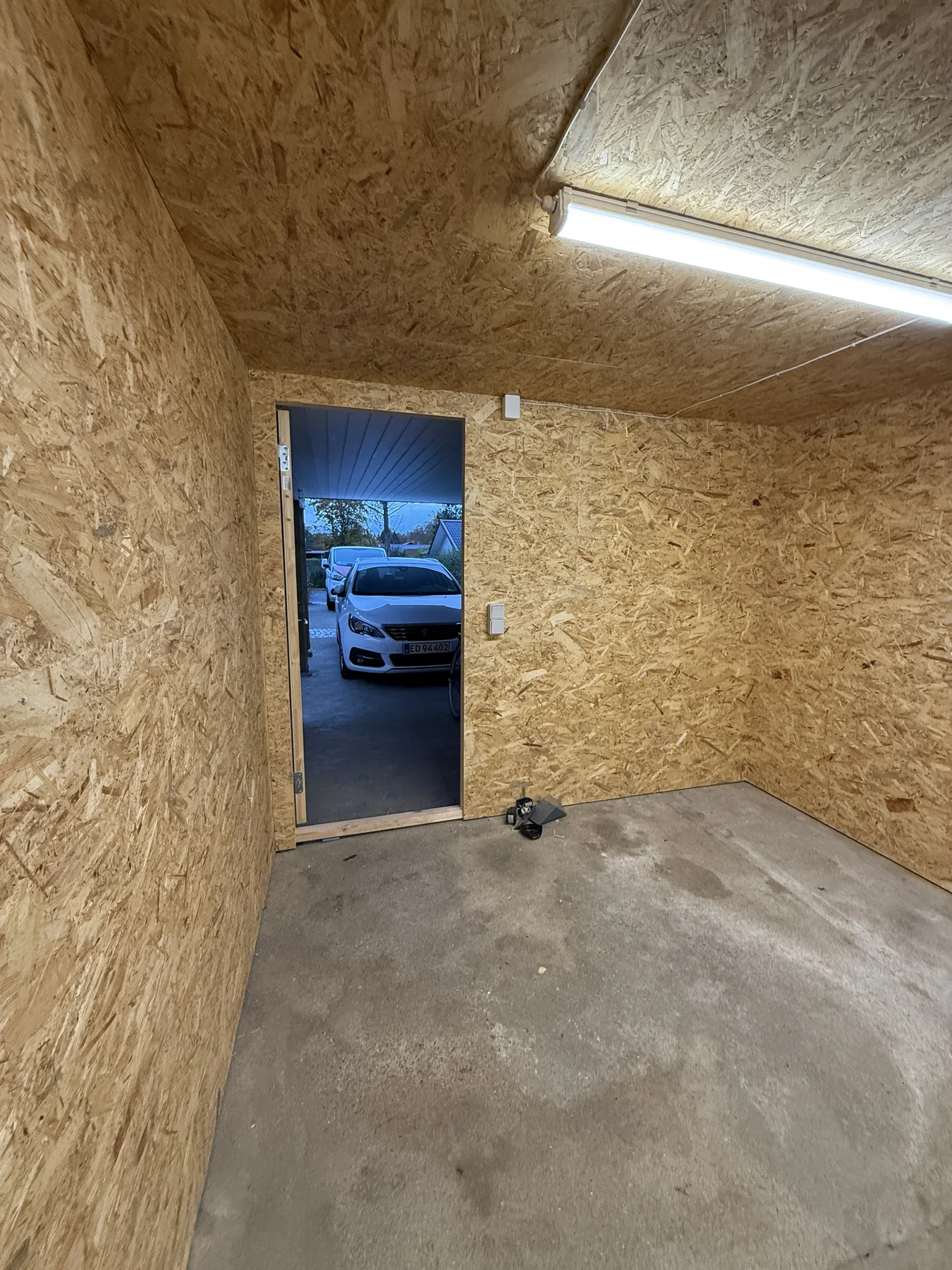 Et tomt rum med OSB-plader på vægge og loft, betongulv, åben dør til hvide biler og en boremaskine ved døren.