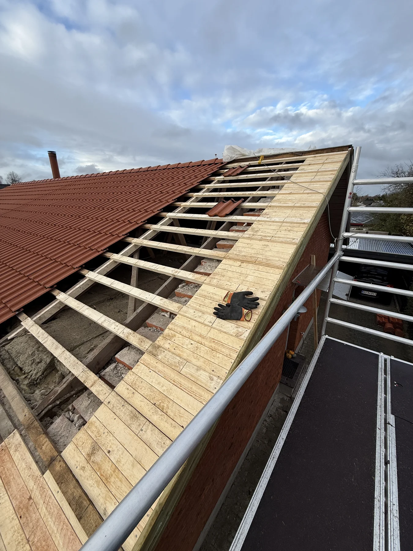 En del af et skråtag er ved at blive renoveret; de gamle tagsten er blevet fjernet, hvilket har blotlagt træbjælkerne, og nye træplanker monteres med omhyggeligt tømrerarbejde. Et par sorte arbejdshandsker ligger på de nyinstallerede planker – et tegn på det igangværende renoveringsarbejde, der udføres af Tømrerfirmaet Kenn. Der ses stilladser.