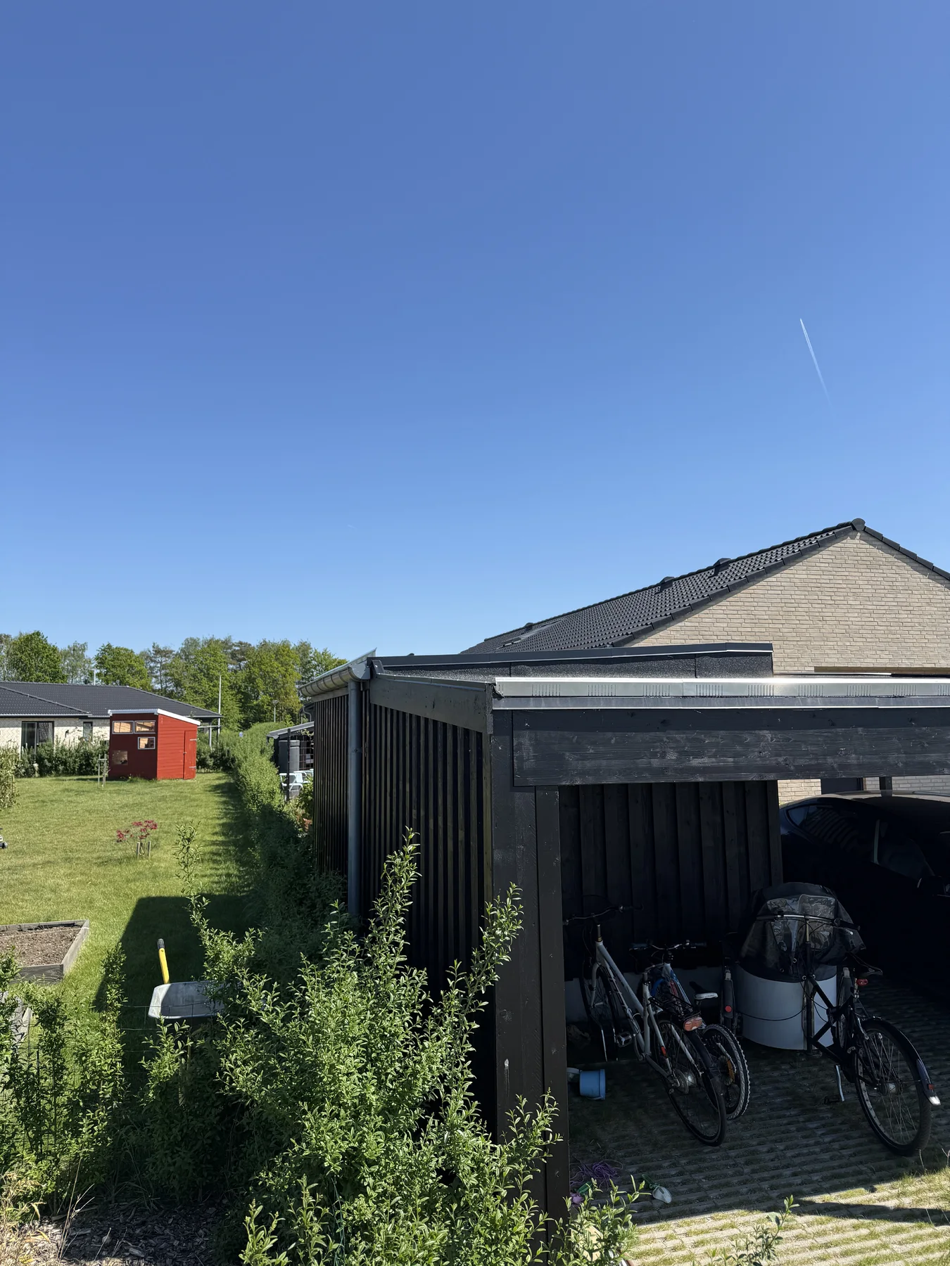 En baghave med en sort carport, hvor der står cykler og diverse genstande, ved siden af et hus, der for nylig er blevet renoveret af Tømrerfirmaet Kenn. Der er en græsplæne, buske, et rødt skur og en klar blå himmel med et synligt kondensspor.