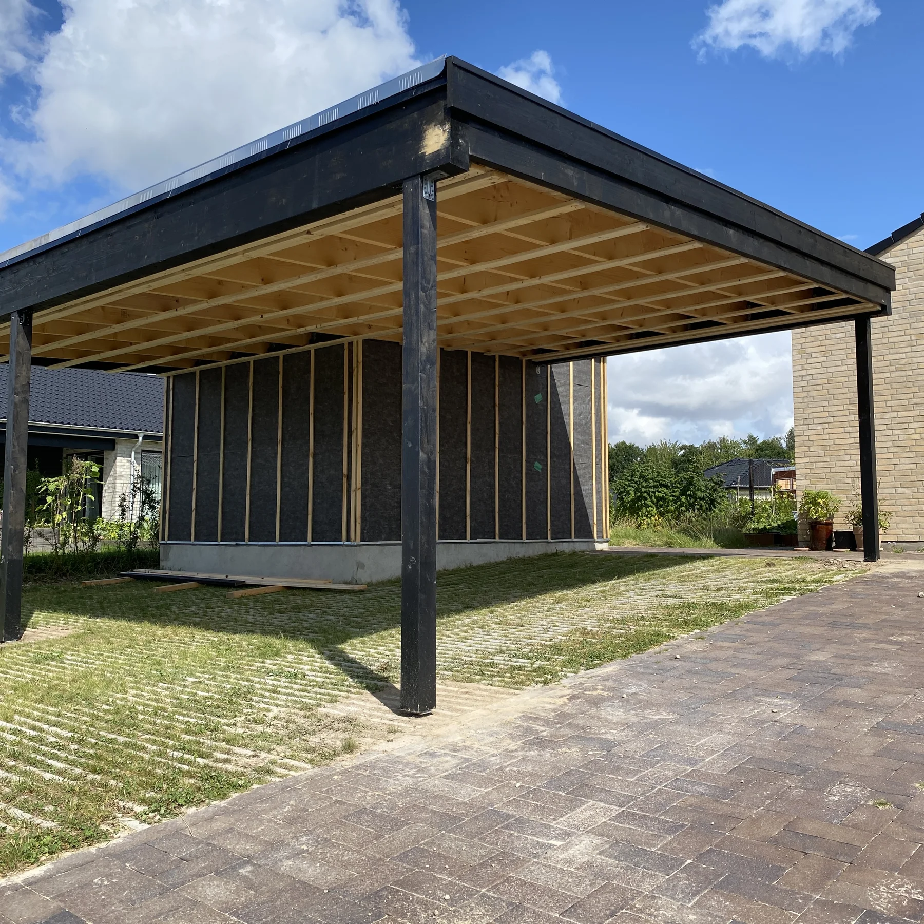 En moderne carport med en åben tagkonstruktion af træ, der er mesterligt udført af Tømrerfirmaet Kenn, står ved siden af en murstensbelagt indkørsel og en delvist færdigbygget bygning. Græs og små planter omgiver området under en delvis overskyet blå himmel, hvilket afspejler den igangværende renovering af huset og det kvalitetsrige tømrerarbejde på stedet.