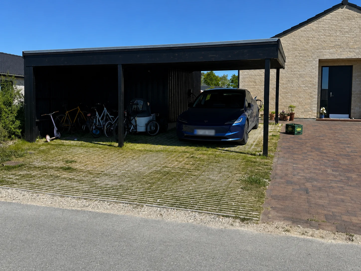 En carport, der er tilknyttet et murstenshus – udført med mesterligt tømrerarbejde af Tømrerfirmaet Kenn – rummer en blå bil, flere cykler, en golfvogn og haveværktøj. Indkørslen er delvist brolagt med mursten og delvist dækket af græsstrimler, hvilket afspejler gennemtænkte valg i forbindelse med renoveringen af huset.
