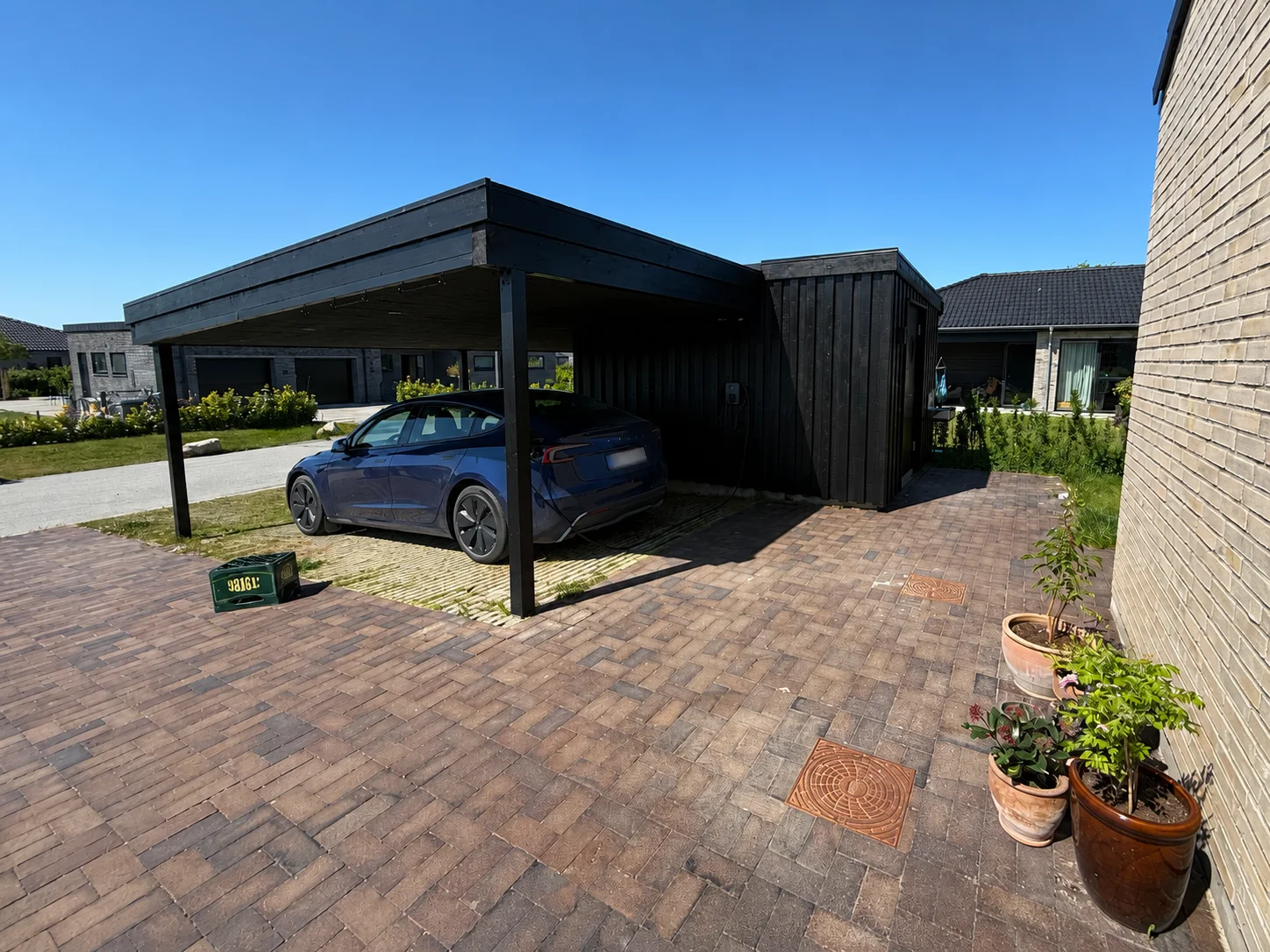En blå bil står parkeret under en sort træcarport – mesterligt udført af dygtige tømrere – ved siden af et beige murstenshus. Krukkeplanter står på række langs kanten af den asfalterede indkørsel og bidrager til den indbydende stemning. Himlen er klar og blå og danner den perfekte kulisse for denne smukke boligrenovering.