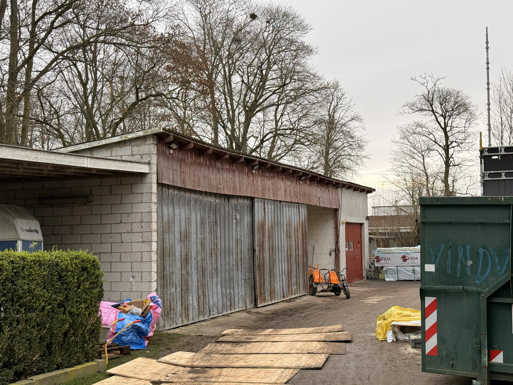 Ved siden af en hæk står en lagerbygning med metaldøre og en betonmur. I forgrunden ses træplanker, en håndvogn, presenninger og en grøn affaldscontainer – tegn på, at Tømrerfirmaet Kenn er i gang med tømrer- eller renoveringsarbejde. I baggrunden ses nøgne træer uden blade.