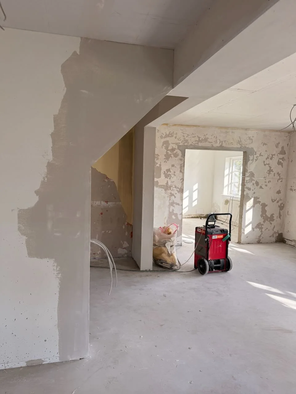 Et rum under renovering med ufærdige vægge, synlige ledninger og spredte byggematerialer afspejler det igangværende arbejde med boligrenovering. Sollys strømmer ind gennem vinduerne og fremhæver det håndværk, der ofte forbindes med tømrerfaget. En rød, transportabel generator eller varmeovn står tæt ved en døråbning – en scene, der minder om projekter, som Tømrerfirmaet Kenn står for.