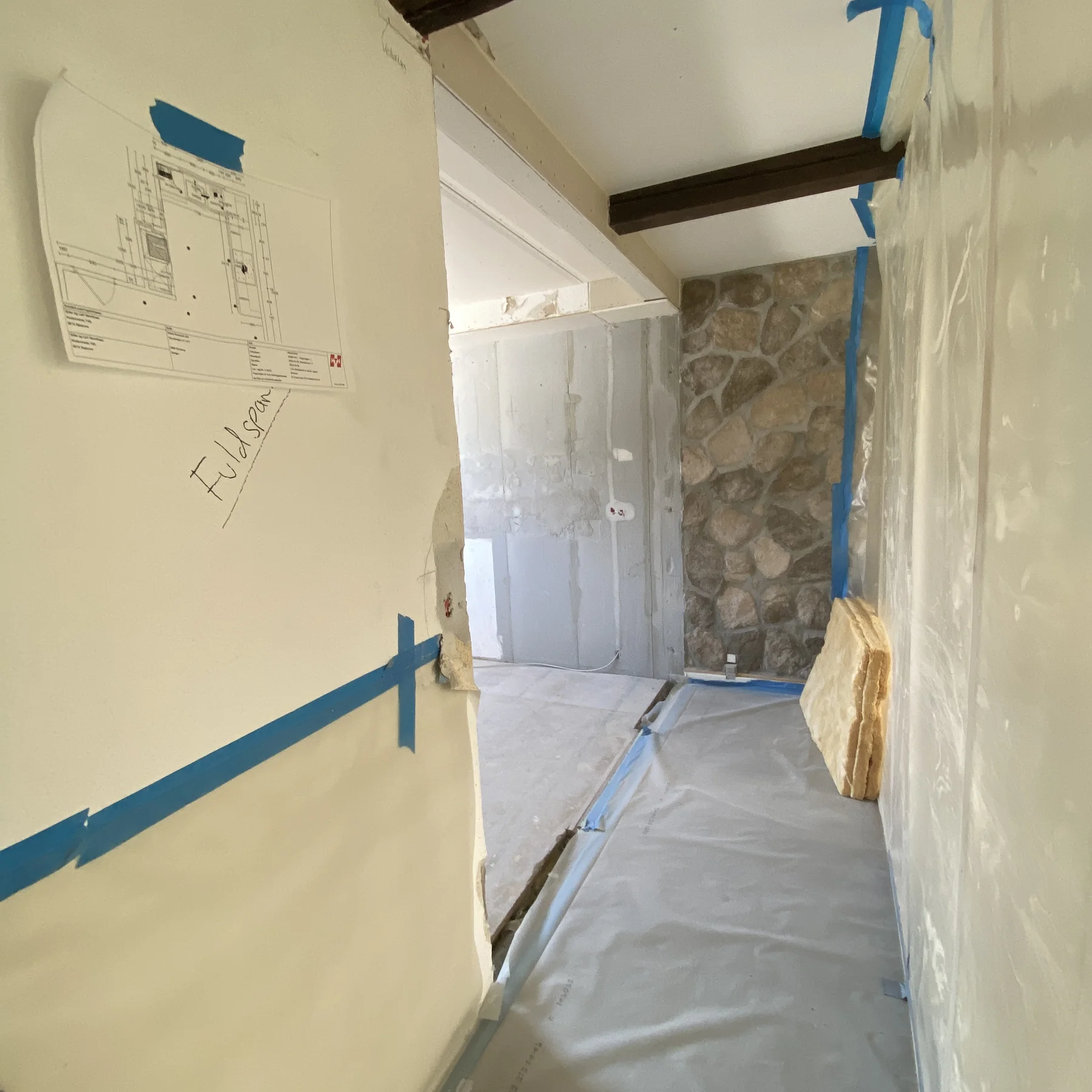En gang, der er under renovering, med plastfolie fastgjort med tape på gulvet og væggene, en stenvæg for enden og en arkitekttegning på papir, der er tapet fast på venstre væg. På højre side ses byggematerialer, der understreger det igangværende tømrerarbejde, som Tømrerfirmaet Kenn udfører som led i dette boligrenoveringsprojekt.
